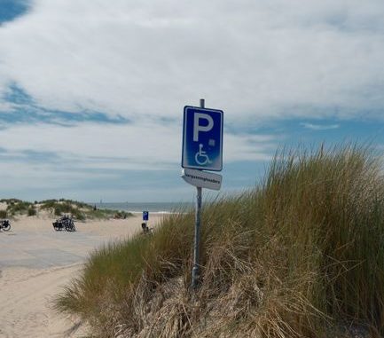 A blue handicap parking sign on a sandy beach with dune grass under a clear sky.